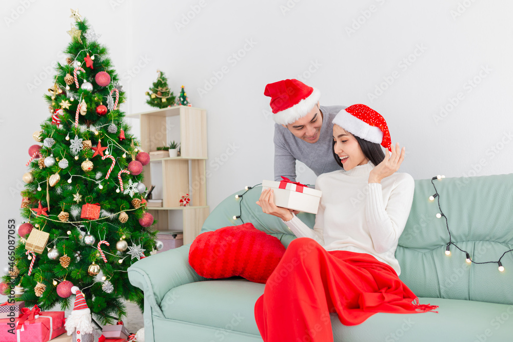 Fototapeta premium asian man send gift box to his girlfriend, asian woman rest on sofa, they holding gift box with hands, they feeling happy and smile, celebration in Christmas day