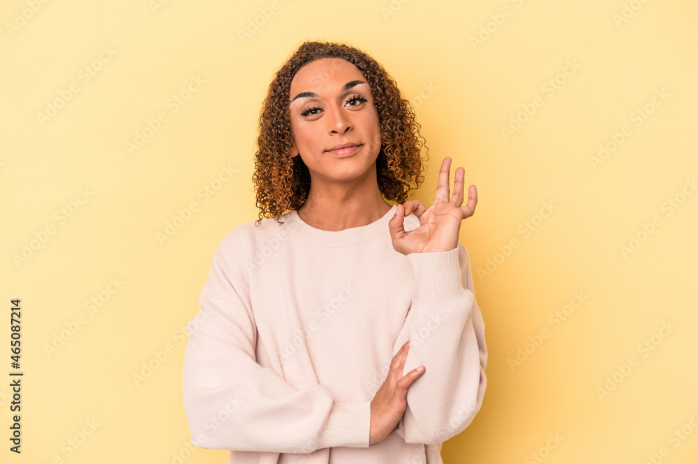 Fototapeta premium Young latin transsexual woman isolated on yellow background winks an eye and holds an okay gesture with hand.