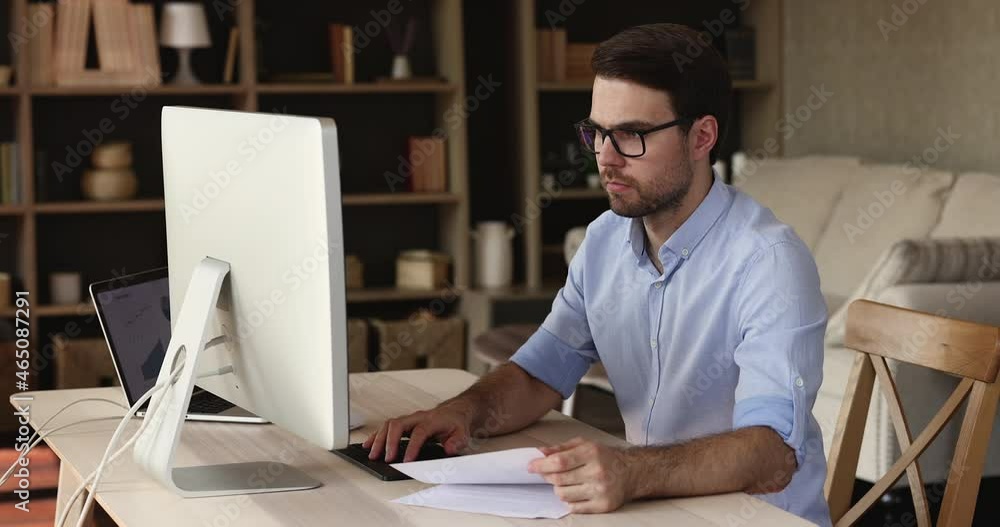 Serious young man marketing specialist work on project from home office using two computers. Professional male social media manager analyze data compare stats information at paper report and online Serious young man marketing specialist work on project from home office using two computers. Professional male social media manager analyze data compare stats information at paper report and online