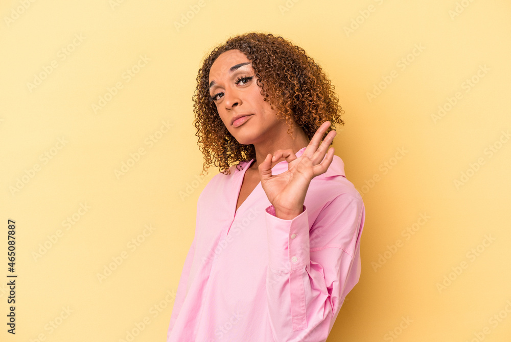 Young latin transsexual woman isolated on yellow background winks an eye and holds an okay gesture with hand. Young latin transsexual woman isolated on yellow background winks an eye and holds an okay gesture with hand.