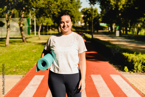 Body positive young caucasian plump fat plus-size woman athlete in fitness outfit preparing before yoga training stretching workout on mat in stadium outdoors