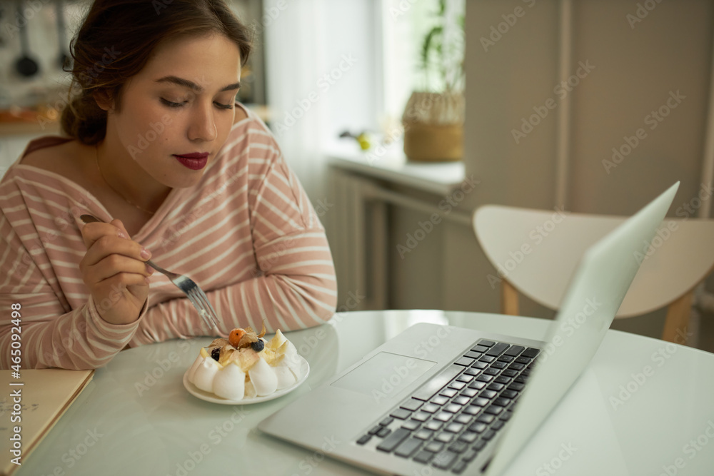 Indoor portrait of beautiful plus-size female teenager with bright make-up watching series on laptop and looking at lovely tasty cake, holding fork, ready to eat it. Bad habits, obesity