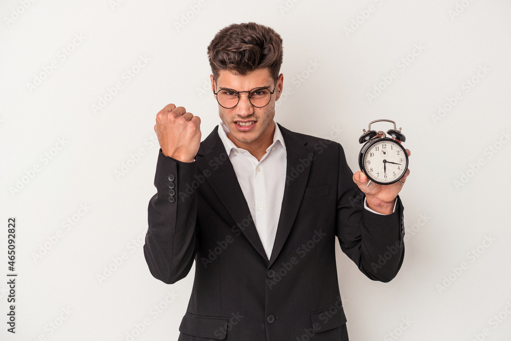 Young business caucasian man holding alarm clock isolated on white background showing fist to camera, aggressive facial expression. Young business caucasian man holding alarm clock isolated on white background showing fist to camera, aggressive facial expression.