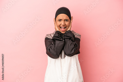 Young Arab woman with sport burqa isolated on pink background covering ears with hands trying not to hear too loud sound.