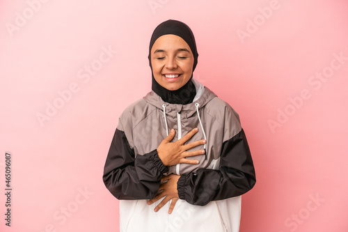 Young Arab woman with sport burqa isolated on pink background laughs happily and has fun keeping hands on stomach.
