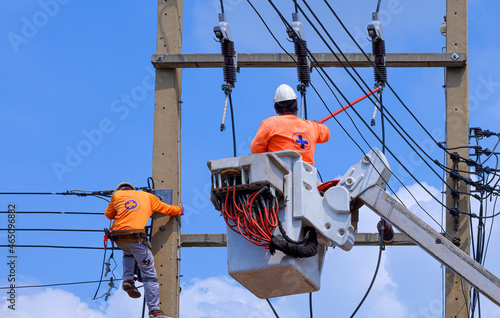 Rear view of 2 electricians with articulated boom lift and disconnect stick tool are working to install electrical transmission on power pole against blue sky background