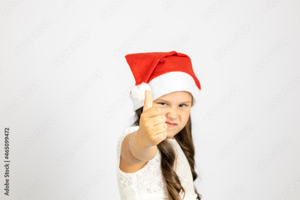 portrait of girl in white dress and wearing red Santa Claus hat gives thumbs up, isolated on white background with copy space. portrait of girl in white dress and wearing red Santa Claus hat gives thumbs up, isolated on white background with copy space.