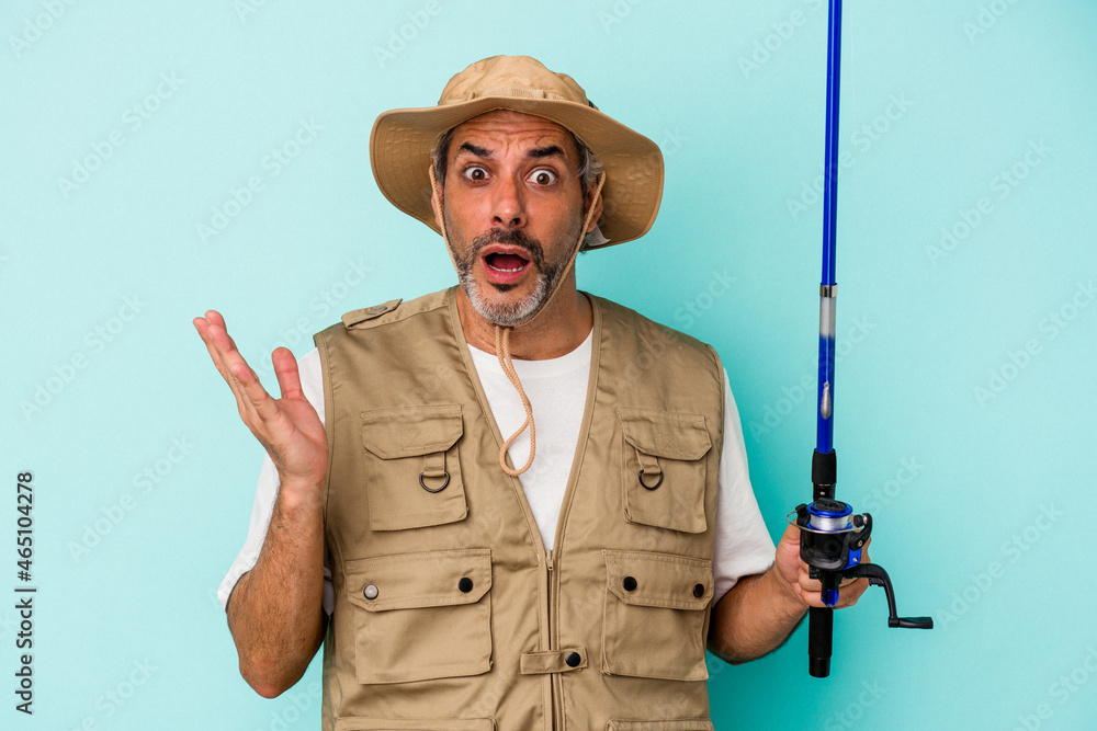 Middle age caucasian fisherman holding rod isolated on blue background surprised and shocked. Middle age caucasian fisherman holding rod isolated on blue background surprised and shocked.