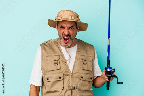 Middle age caucasian fisherman holding rod isolated on blue background screaming very angry and aggressive.