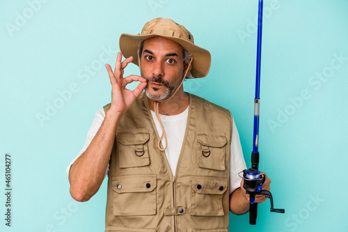 Middle age caucasian fisherman holding rod isolated on blue background with fingers on lips keeping a secret.