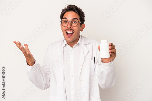 Young pharmacist mixed race man holding pills isolated on white background receiving a pleasant surprise, excited and raising hands.