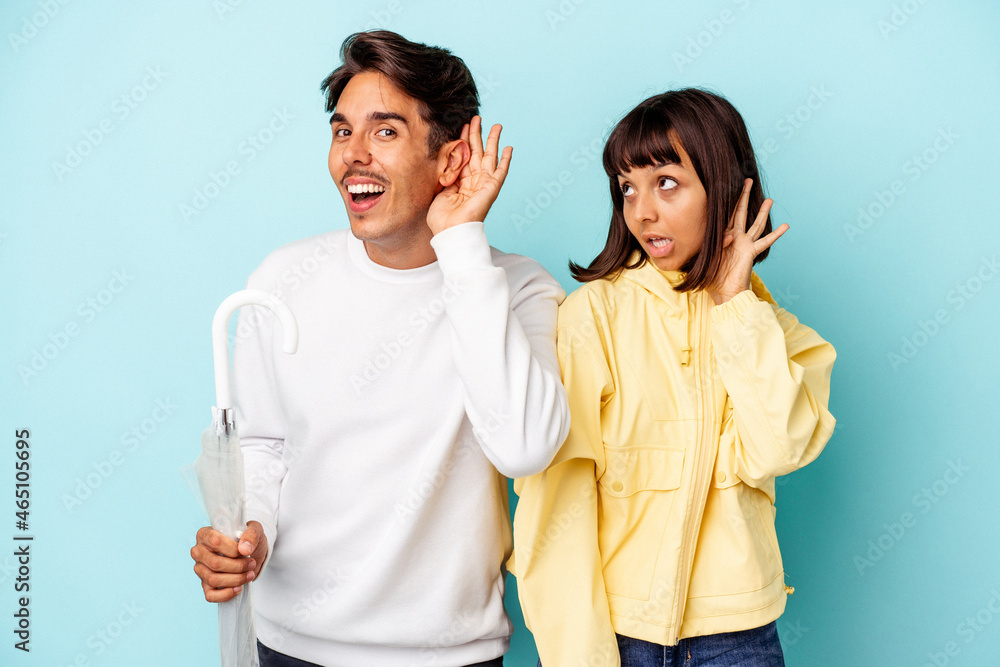 Young mixed race couple holding umbrella isolated on blue background trying to listening a gossip. Young mixed race couple holding umbrella isolated on blue background trying to listening a gossip.