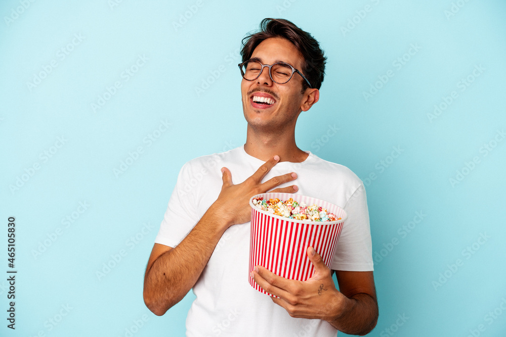 Young mixed race man eating popcorns isolated on blue background laughs out loudly keeping hand on chest.