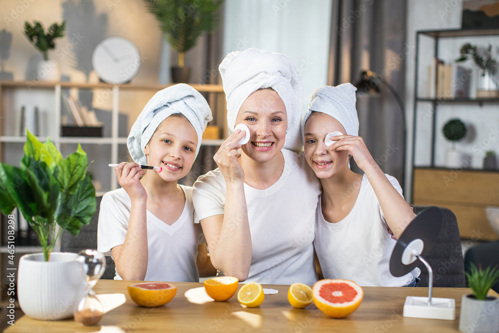 Mother with two daughters cleaning face with cotton pads and applying moustirising creme with brush. Caucasian family of three wearing white t-shirts and bath towel on head. Skin care concept.
