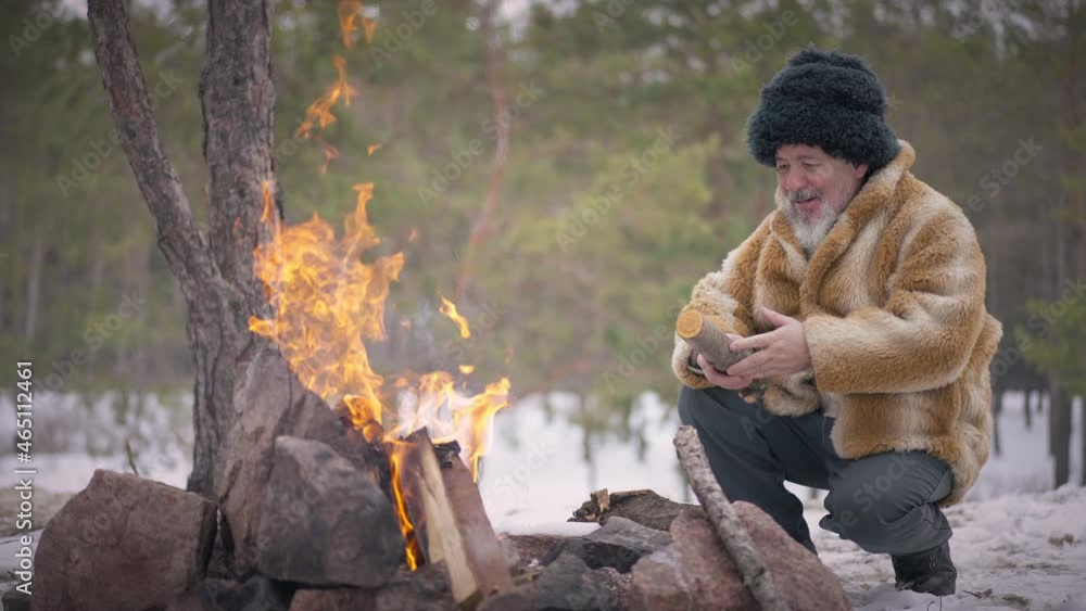 Smiling indigenous old man sitting in winter forest at fire with firewood. Wide shot portrait of confident senior Mongol guy outdoors on cold day. Slow motion Smiling indigenous old man sitting in winter forest at fire with firewood. Wide shot portrait of confident senior Mongol guy outdoors on cold day. Slow motion