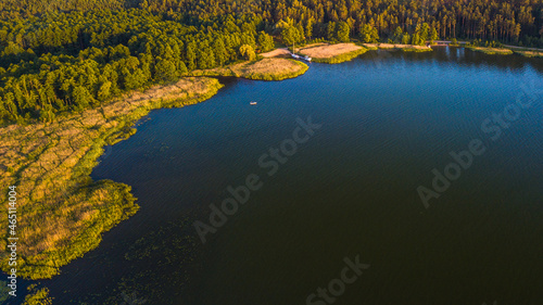 Aerial view of the lake and green forest in a sunny day. Shore of the lake. Small boat sailing on the lake, and the other boats docked in the distance.