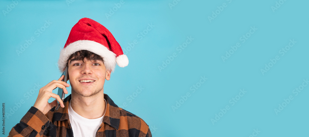 Obraz premium portrait of teenage boy talking on mobile phone wearing santa claus hat isolated on blue background