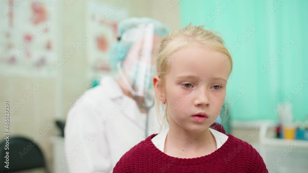 Zoom out rack focus shot of senior doctor in protective face shield and mask listening with stethoscope to lung sound of kid girl breathing deeply in clinic. Patient recovering from covid-19 pneumonia Zoom out rack focus shot of senior doctor in protective face shield and mask listening with stethoscope to lung sound of kid girl breathing deeply in clinic. Patient recovering from covid-19 pneumonia