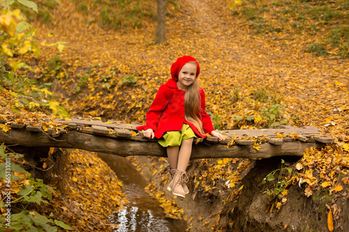 Fototapeta Naklejka Na Ścianę i Meble -  a beautiful happy little blonde girl in a red coat and a red beret is sitting on a bridge across a river in the city's autumn yellow park