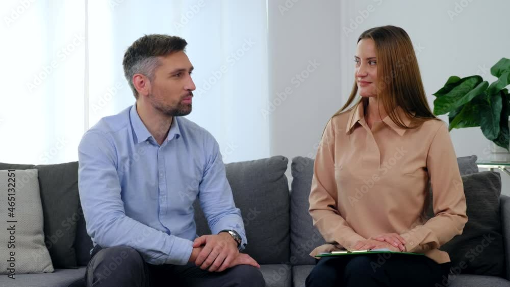 Man patient and woman psychologist sitting on couch talking and turn to camera after having passed psychotherapy session in medical hospital clinic office. Mental health problems after coronavirus Man patient and woman psychologist sitting on couch talking and turn to camera after having passed psychotherapy session in medical hospital clinic office. Mental health problems after coronavirus