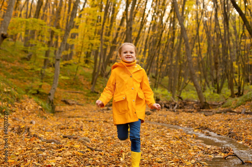 a beautiful happy little blonde girl in a yellow raincoat and yellow boots runs near the river in the urban autumn forest