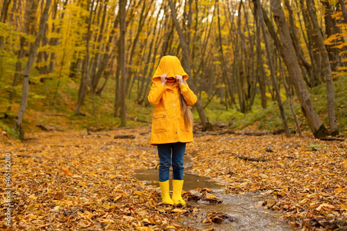 a beautiful happy little blonde girl in a yellow raincoat and yellow boots jumps in the river that flows through the city forest autumn © Тамара Киреева