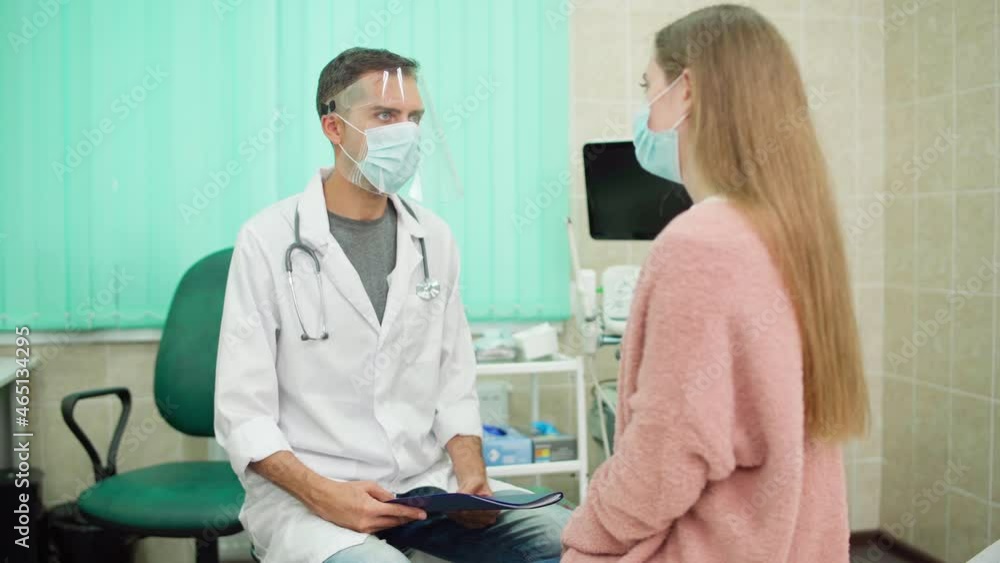 Doctor in protective face shield holding medical history talking to female patient in face mask in clinic. Young male physician in white coat working with coronavirus covid-19 patients