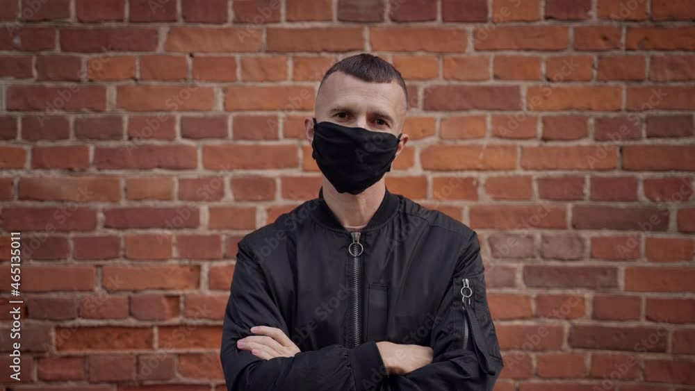 A middle-aged short haired man in a black jacket with the appearance of a gopnik against the background of a brick wall removes a protective mask, spits, looks dissatisfied at the camera. A middle-aged short haired man in a black jacket with the appearance of a gopnik against the background of a brick wall removes a protective mask, spits, looks dissatisfied at the camera.