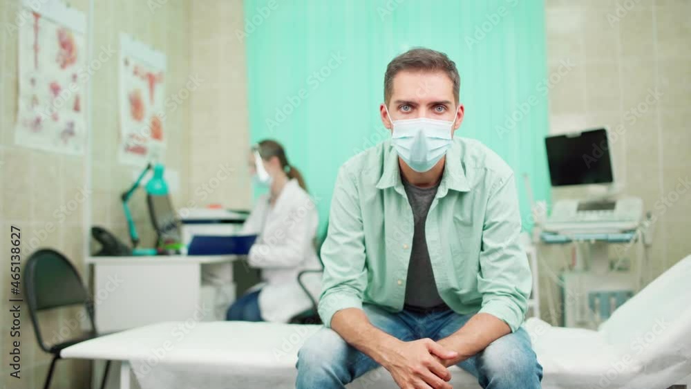 Sad male patient in protective face mask thinking and looking at camera sitting on couch in doctors office waiting for physician making notes in medical history. Sick man visiting clinic in pandemic Sad male patient in protective face mask thinking and looking at camera sitting on couch in doctors office waiting for physician making notes in medical history. Sick man visiting clinic in pandemic