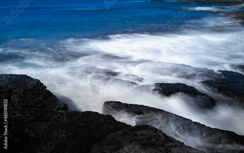 Fototapeta Naklejka Na Ścianę i Meble -  Smooth silky waves flow over the black rocks on the beach. Panoramic motion blur rocky shore image.