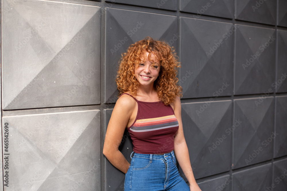 Fototapeta premium young redhead with curly hair and jeans smiling in the city with a gray wall in the background