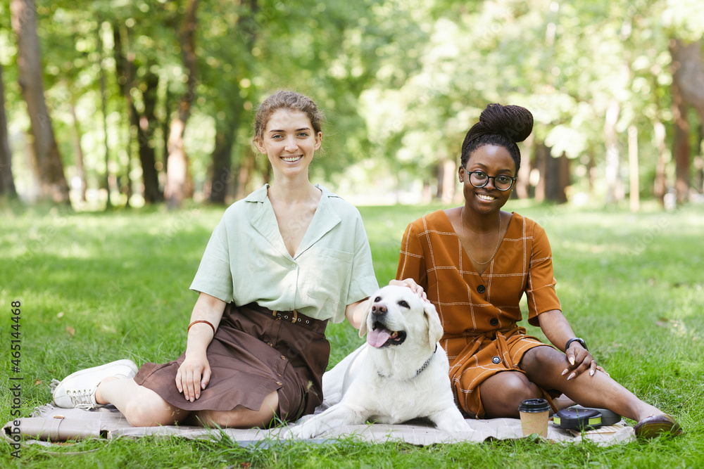 Full length portrait of two young women sitting on grass in park with dog outdoors and looking at camera, copy space Full length portrait of two young women sitting on grass in park with dog outdoors and looking at camera, copy space