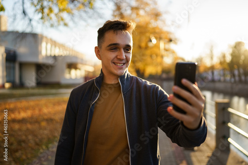 Young caucasian man standing outdoor alone in autumn evening making a phone video call or selfie photo smiling happy