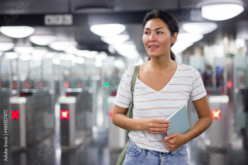 Asian woman standing in subway station with ticket gates in background.