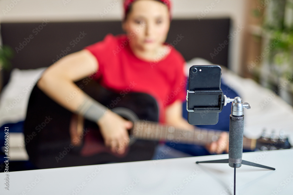 Close up shot of a smartphone on a tripod. Young woman recording herself playing acoustic guitar while spending time at home Close up shot of a smartphone on a tripod. Young woman recording herself playing acoustic guitar while spending time at home