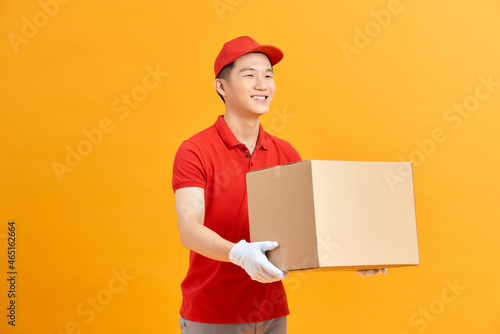 Image of a happy young delivery man in red cap standing with parcel post box isolated over yellow background.