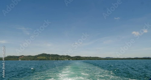 The ferry is sailing out into the ocean. There was a wave behind the boat. There is a coast and hills in front. and the blue sky