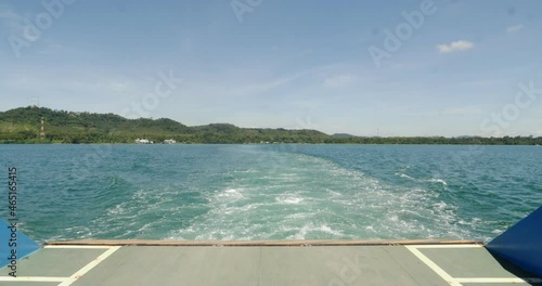 The ferry is sailing out into the ocean. There was a wave behind the boat. There is a coast and hills in front. and the blue sky