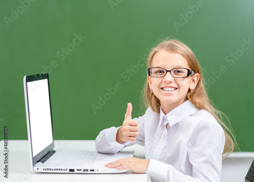 Happy smart girl uses laptop near the green school chalkboard and shows thumbs up gesture