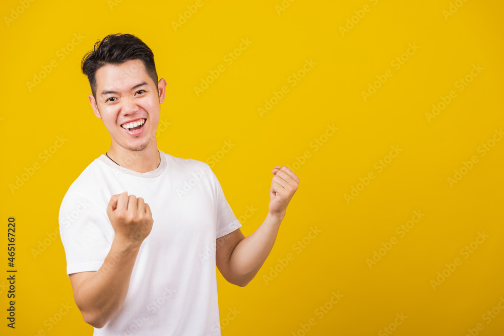 Asian handsome young man smiling positive shaking hands enthusiastic shouting yes for win competition, male raising his fists with smiling delighted face, studio shot isolated on yellow background Asian handsome young man smiling positive shaking hands enthusiastic shouting yes for win competition, male raising his fists with smiling delighted face, studio shot isolated on yellow background