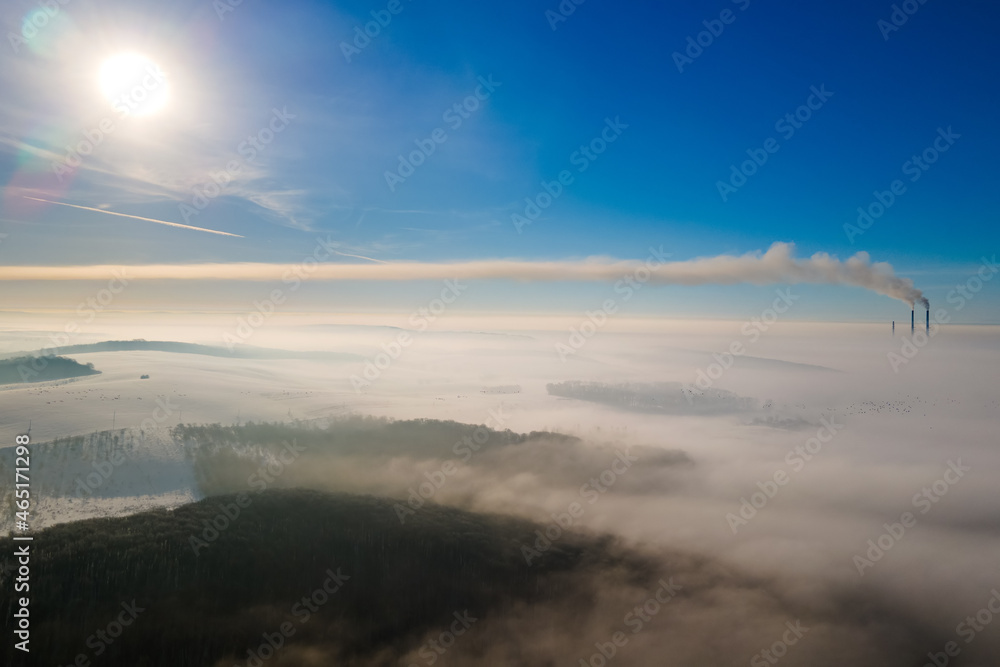 Aerial view of winter landscape with foggy countryside and distant factory pipes emmiting black dirty smoke polluting environment.