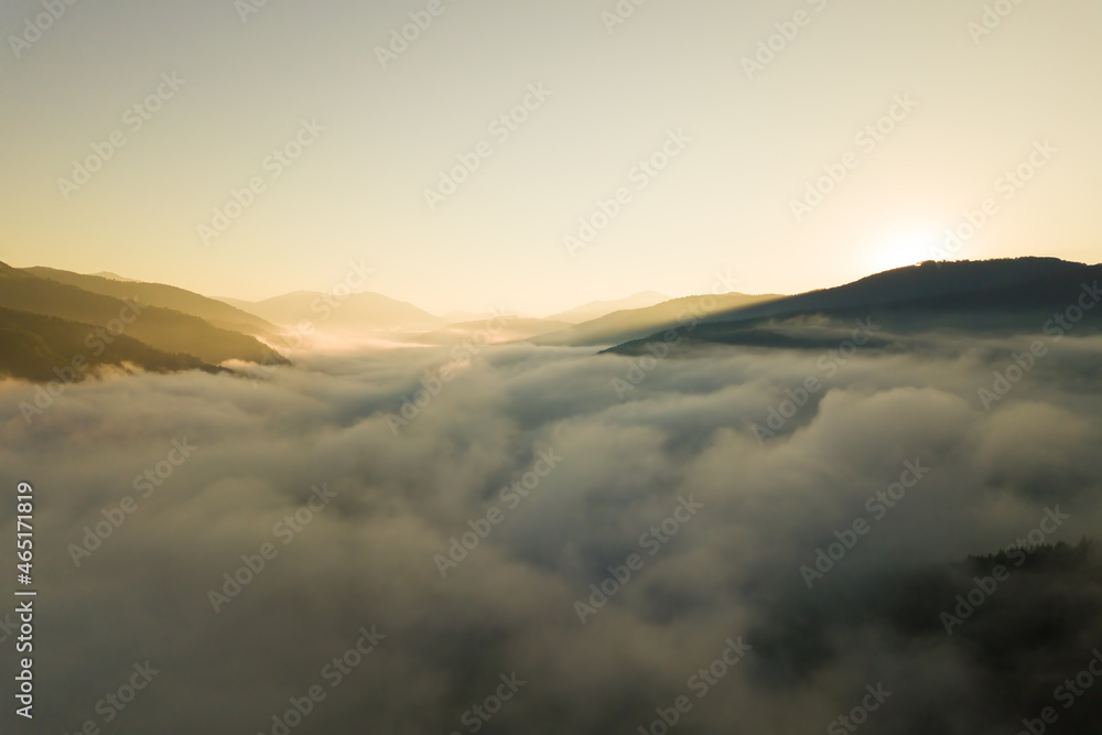 Aerial view of vibrant sunrise over white dense fog with distant dark silhouettes of mountain hills on horizon.