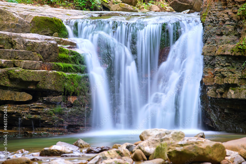 Waterfall on mountain river with white foamy water falling down from rocky formation in summer forest Waterfall on mountain river with white foamy water falling down from rocky formation in summer forest