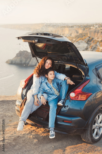 A young mother and her teenage son in denim enjoy the view of the sea and mountains on the top of the hill, sitting in the open trunk of a car and hugging together. Happy family relationship concept. © Evgeniya Grande