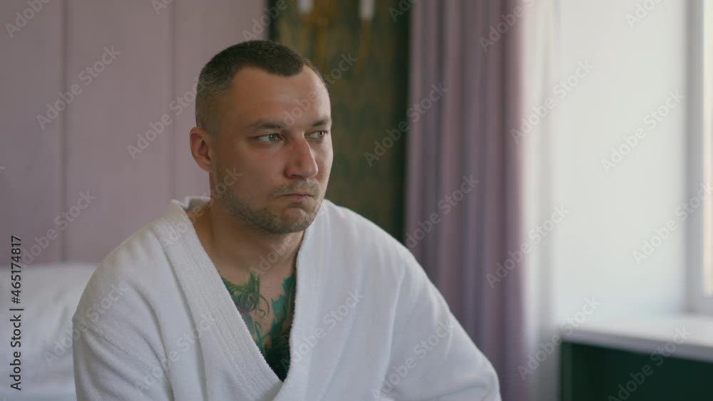 Thoughtful man guest with tattoos on chest in terry bathrobe sits on bed in comfortable hotel room in morning close view slow motion Thoughtful man guest with tattoos on chest in terry bathrobe sits on bed in comfortable hotel room in morning close view slow motion