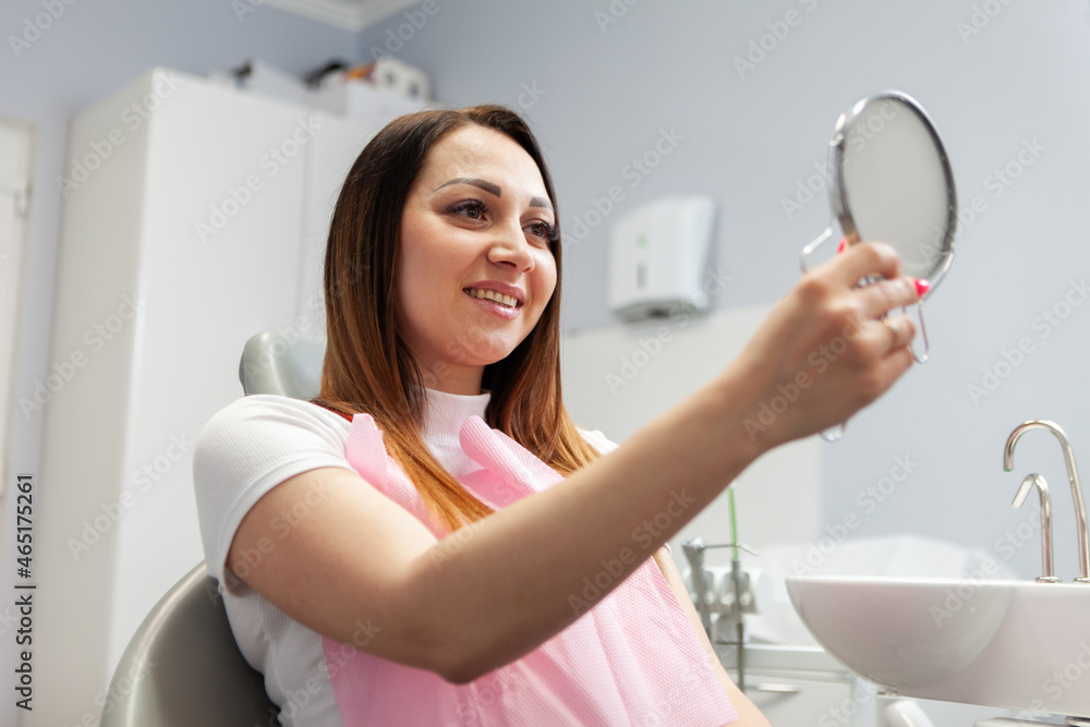 Happy woman looks at her smile in the mirror on visit to the dentist in dental office. Beautiful smile, whitening, dental treatment concept