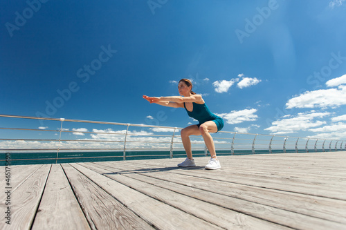 Fit woman doing squat exercise, training legs on the embankment at bright sunny day with blue sky and clouds