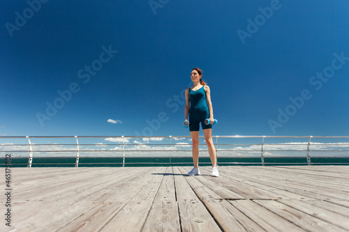 Sporty Caucasian woman exercising with dumbbells on the embankment at bright sunny day with blue sky and clouds. Healthy lifestyle concept. Wide angle