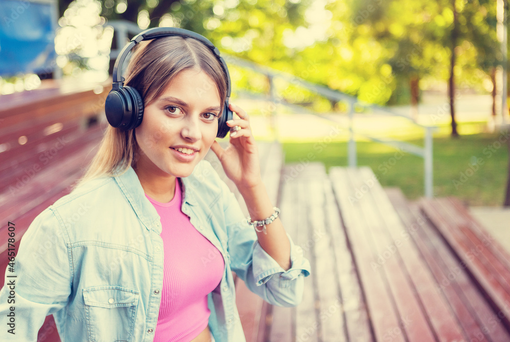 Image of a young stylish woman in youth fashionable clothes listening to music with headphones while sitting in the stands outdoors Image of a young stylish woman in youth fashionable clothes listening to music with headphones while sitting in the stands outdoors