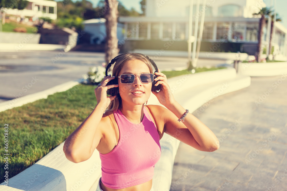 Beauty blonde woman dressed in stylish clothes and sunglasses listen music with headphones outdoors at summer bright sunny day